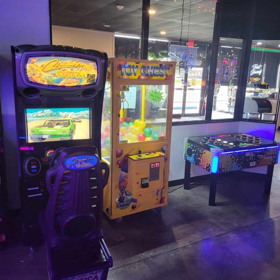 Arcade games, including a racing simulator, claw machine, and air hockey table, in a restaurant.