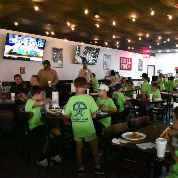 Children in green shirts eating pizza at tables in Decades Family Restaurant, with TVs and "ROLL TIDE" banner.