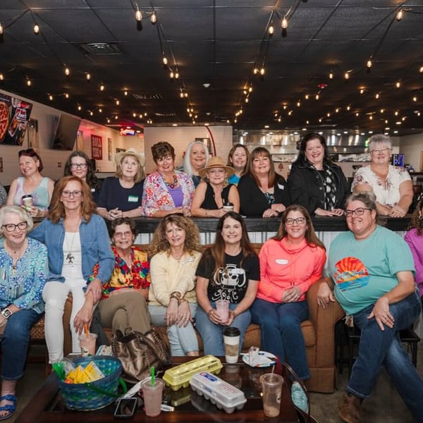 Group of women smiling at Decades Family Restaurant, with string lights and pizza signs in the background.