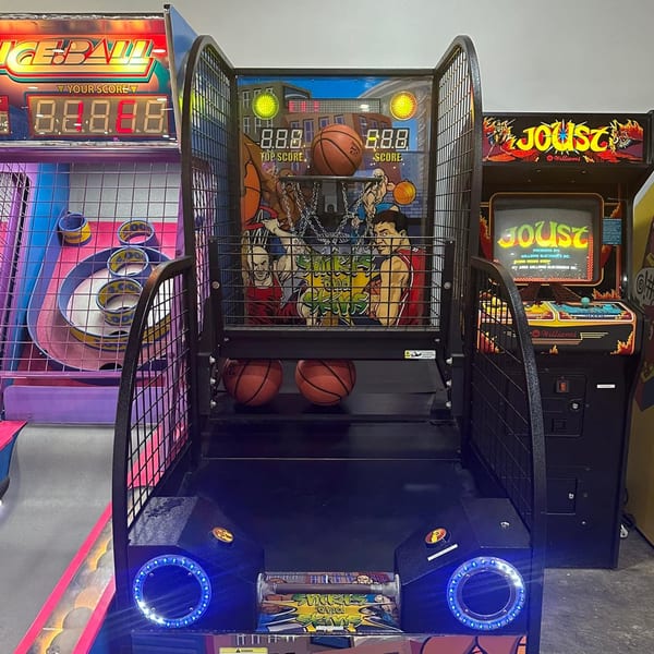 A basketball arcade game with two basketballs, next to a skeeball game and a Joust arcade machine.