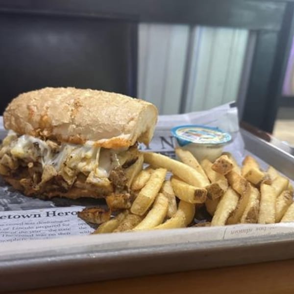 Cheesesteak sandwich with melted cheese, a side of fries, and a dipping sauce on a metal tray.