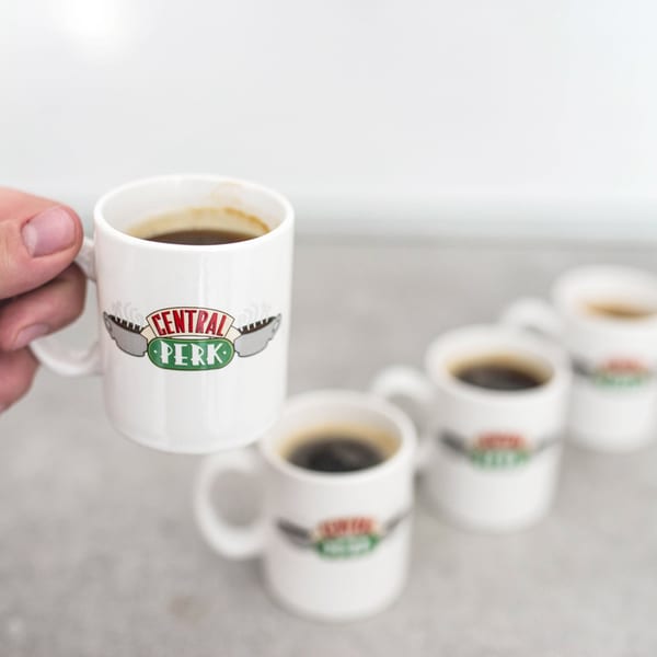 A hand holds a white Central Perk coffee mug, with several more blurred in the background.