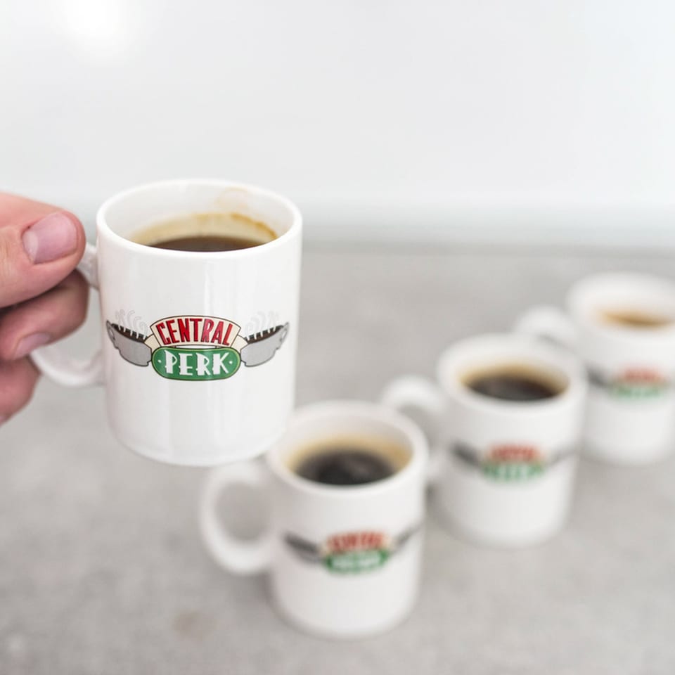 A hand holds a white Central Perk coffee mug, with several more blurred in the background.