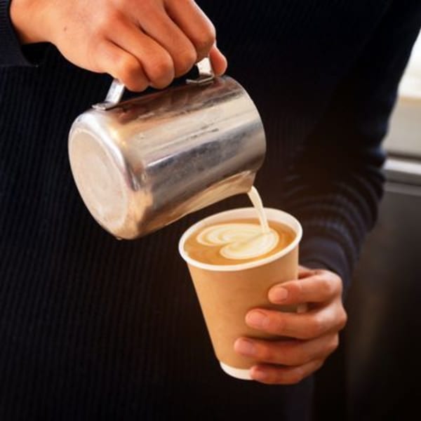 Hands pouring steamed milk from a metal pitcher into a paper cup of coffee, creating latte art.