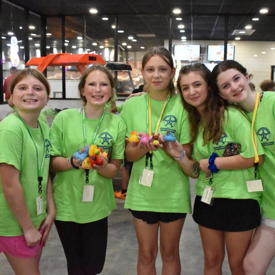 Five smiling girls in green "Shining Star Camp" t-shirts hold colorful rubber toys inside Decades Family Restaurant.