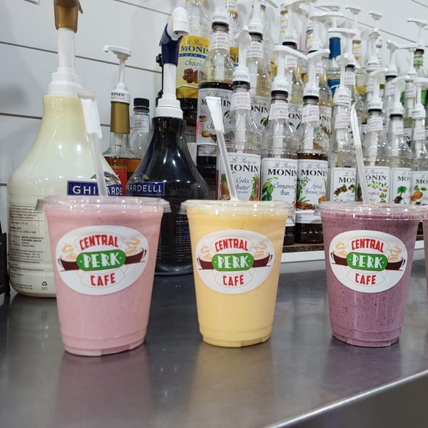 Three Central Perk Cafe smoothies, pink, yellow, and purple, sit on a counter with syrup bottles.