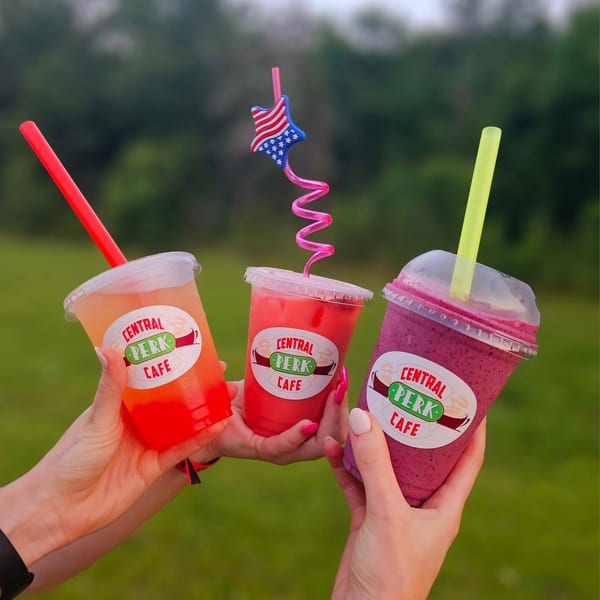 Three hands hold up colorful Central Perk Cafe drinks with a green, blurry background.