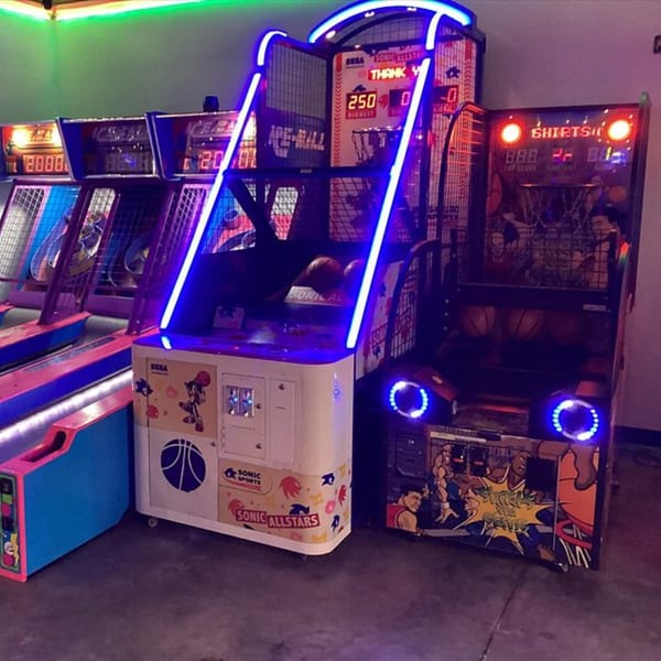 Arcade basketball and Skee-Ball machines with neon lights in a dimly lit game room at Decades Family Restaurant.